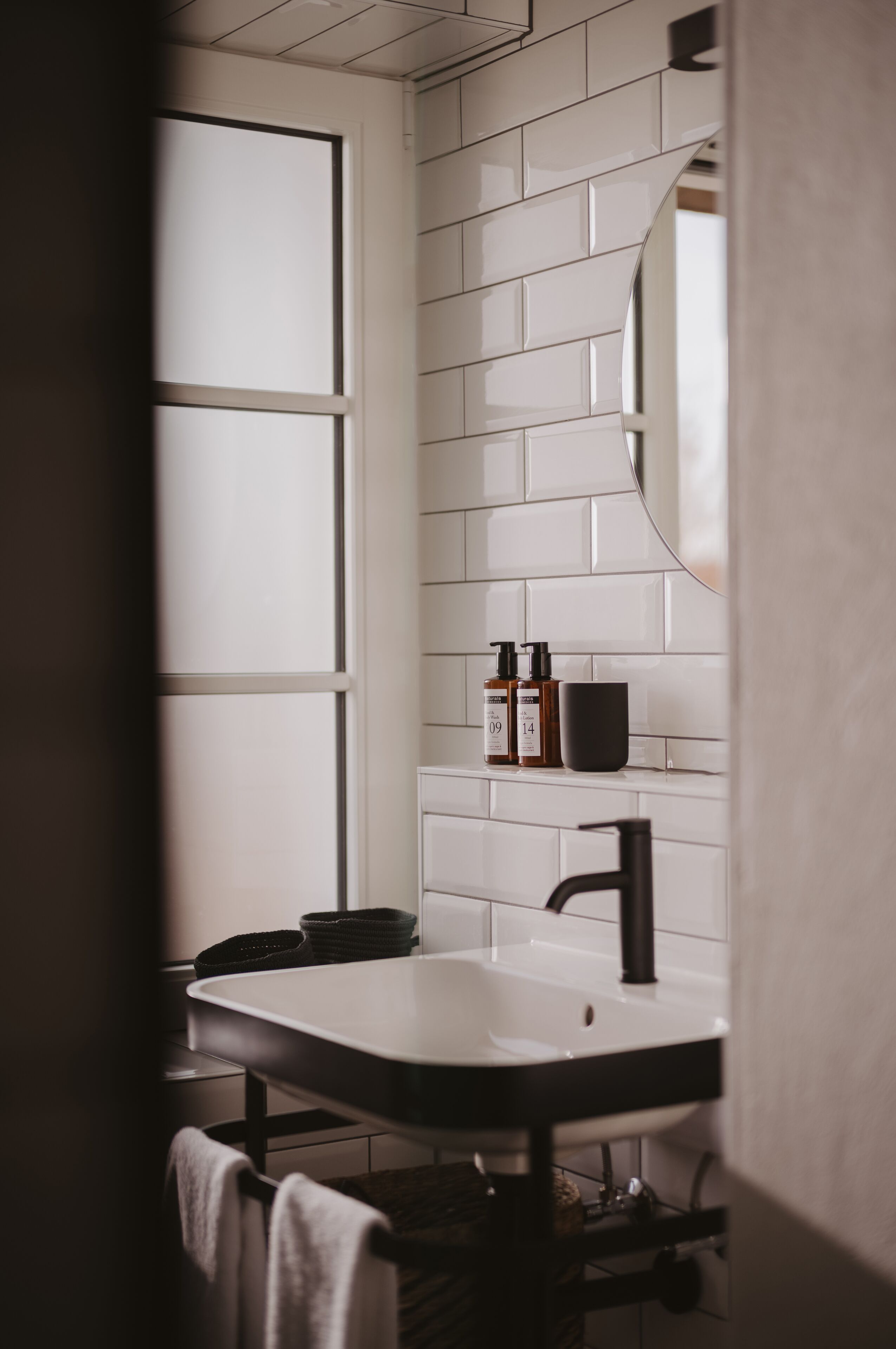 Ein Badezimmer mit weiß gefliesten Wänden, einem schwarzen Waschbecken und einem Spiegel. Zwei Flaschen hängen an der Wand und eine schwarze Tasse steht auf der Ablagefläche.