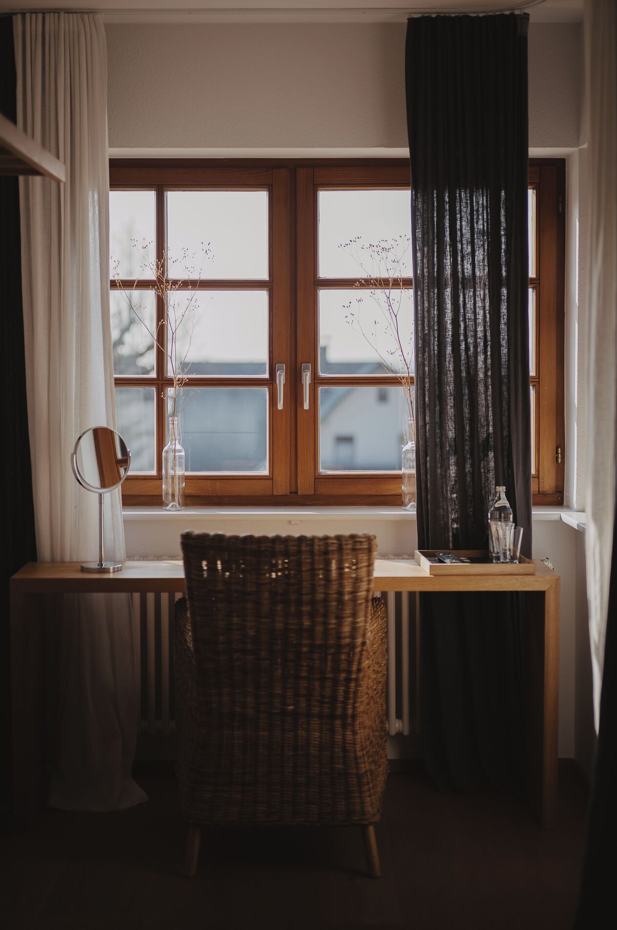 A room with a wooden desk, a wicker chair, a mirror, and a window with curtains.