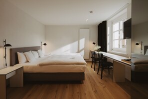 A modern bedroom with a large bed, a desk, and a chair, featuring a window with natural light. - Der Hirschen (Sankt Maergen)