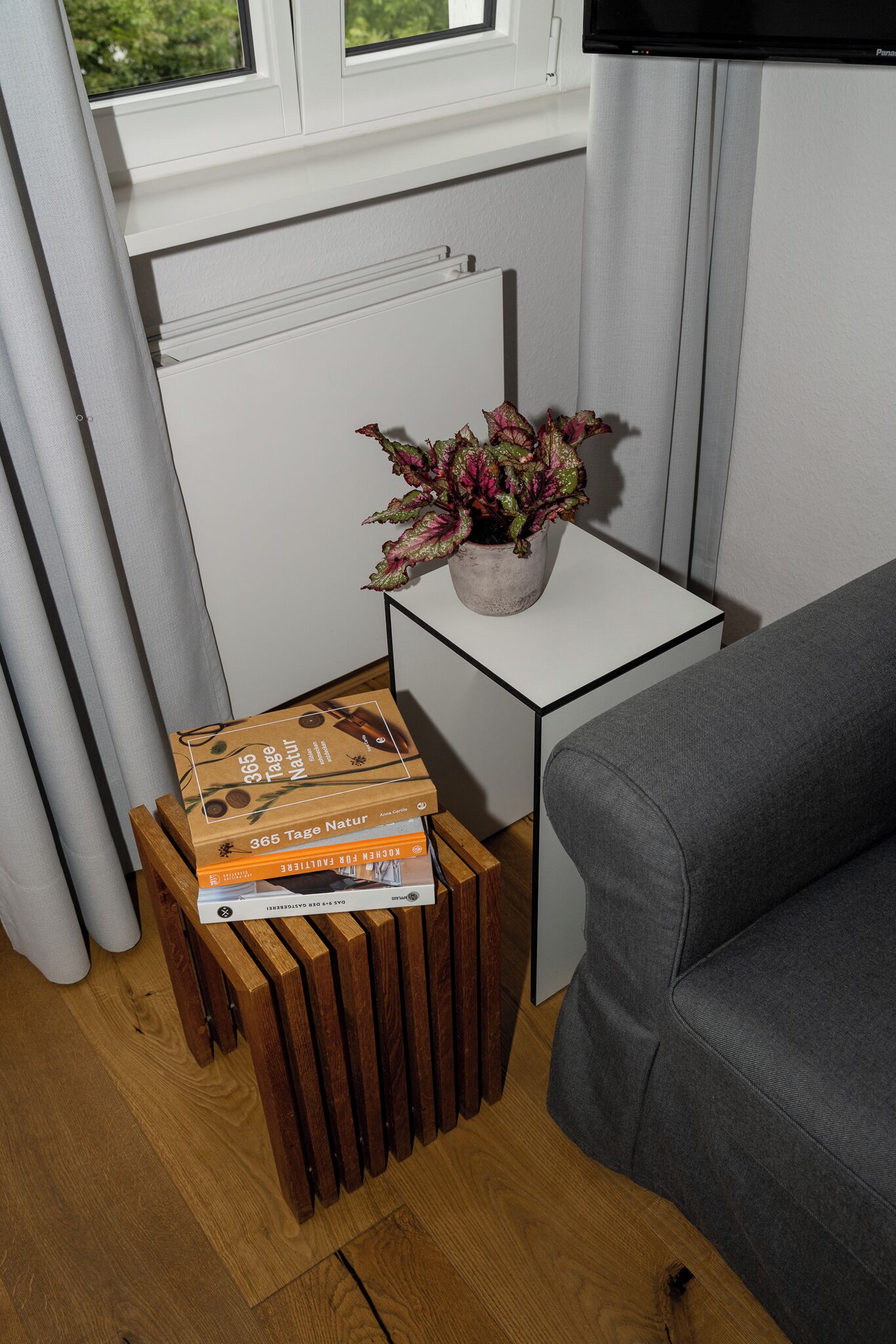 A corner of a room with a grey sofa, a small white table with a potted plant, and a wooden stool with books.