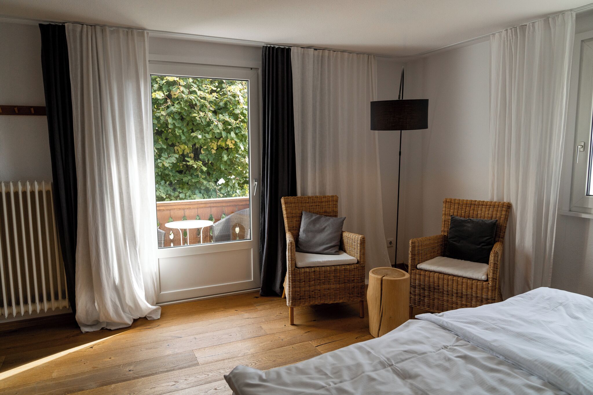 Une chambre d’hôtel avec une grande fenêtre, deux fauteuils en osier avec des coussins, un lit et une lampe sur pied.