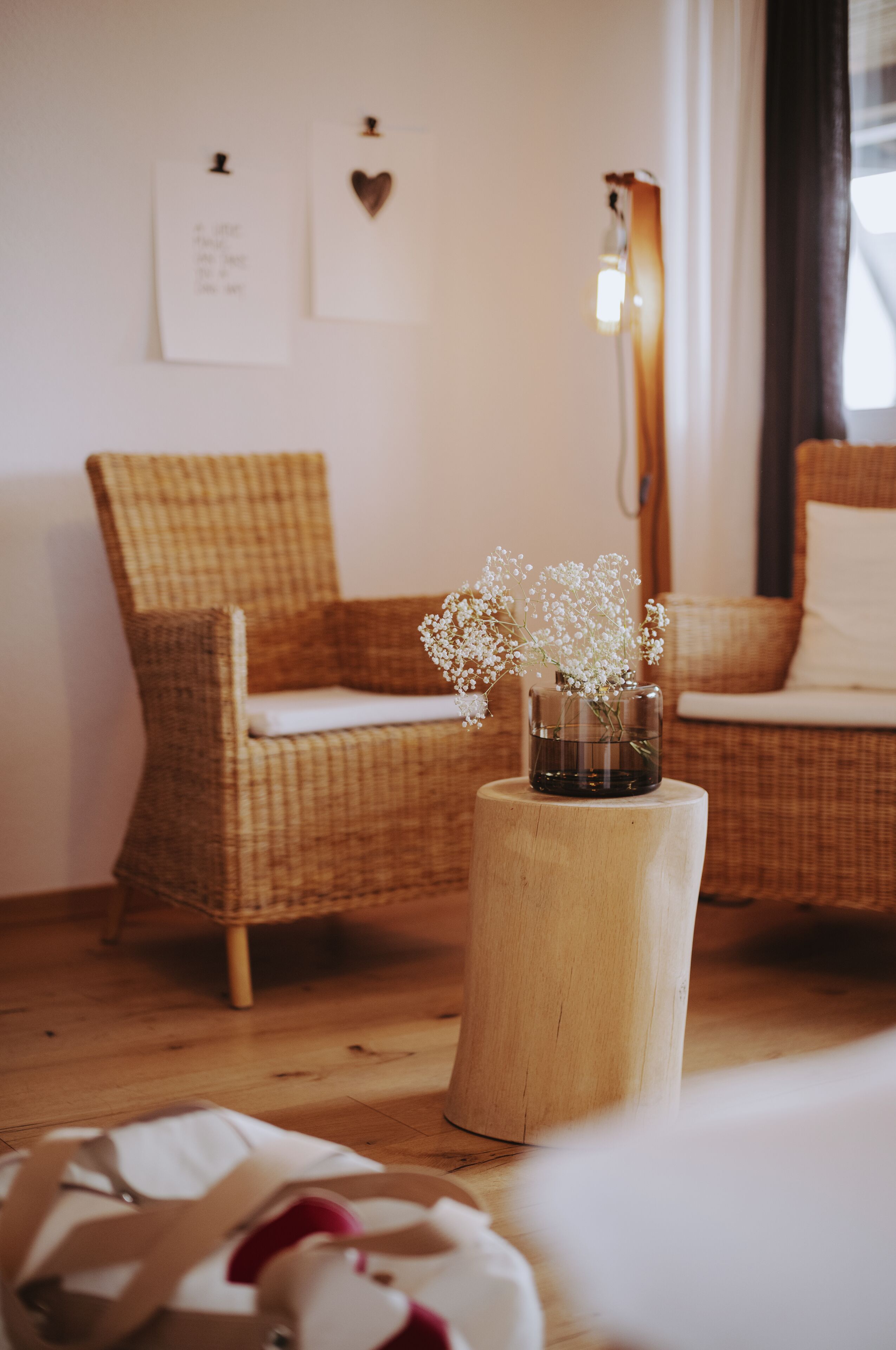 A wicker chair with a cushion, a wooden stool with a glass vase of flowers, and a wall with two framed items.