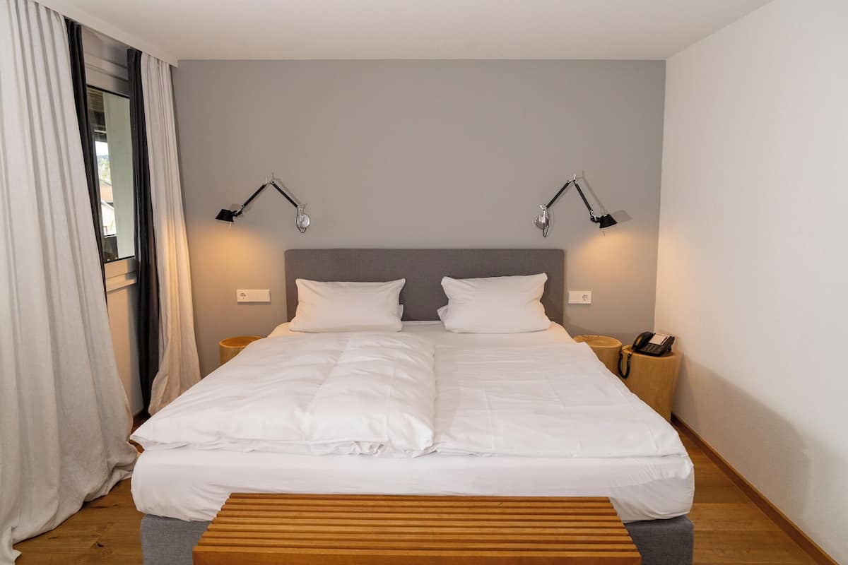 Una cama bien tendida con ropa de cama blanca, dos lámparas de noche, una mesita de noche de madera con un teléfono y una ventana con cortinas.