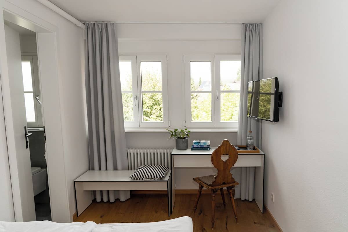 Un dormitorio con cama, escritorio y silla, radiador, ventana con cortinas, televisor y planta.
