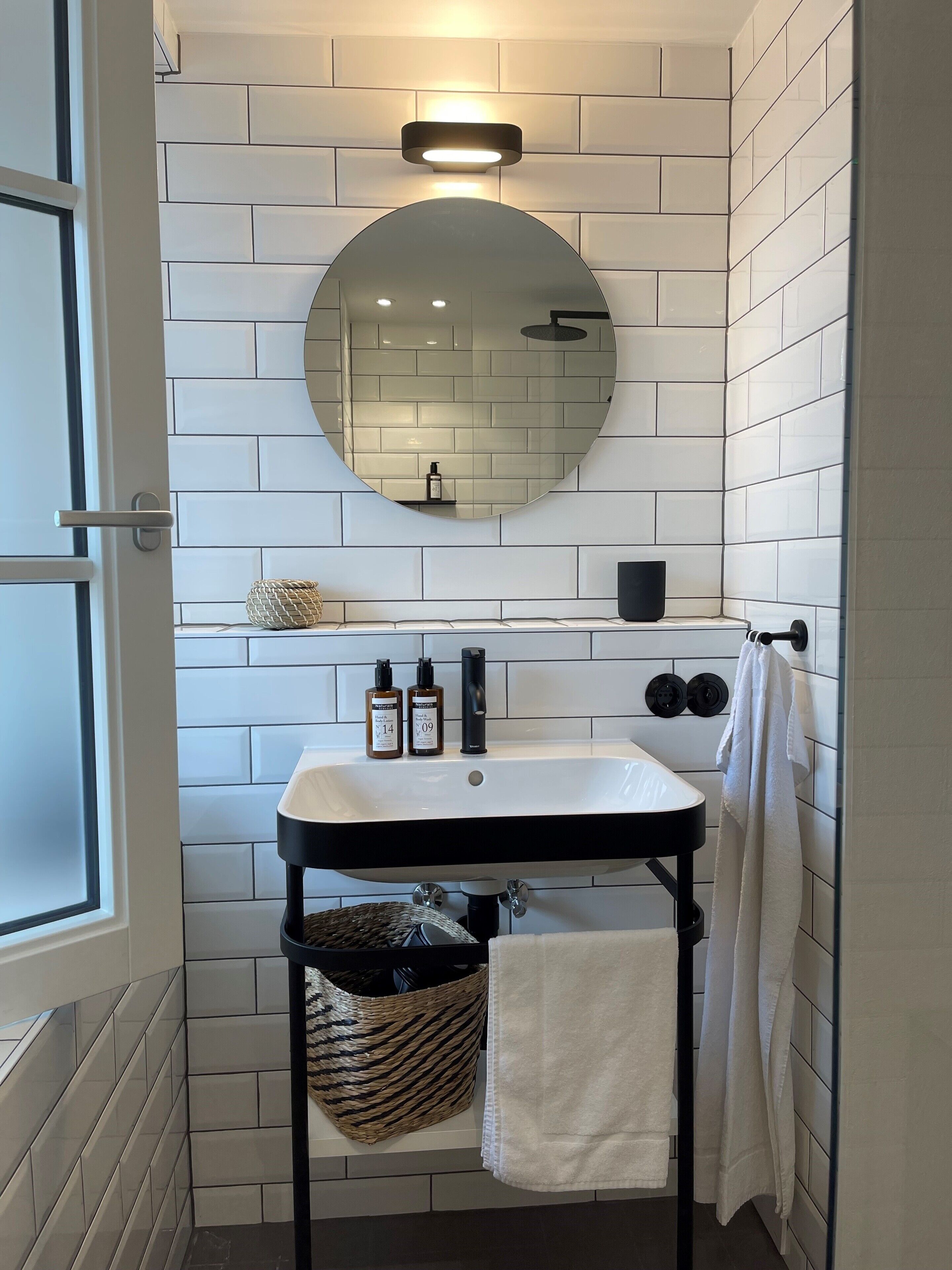 A bathroom with a white sink, black faucet, and a round mirror on the wall. There is a towel rack with a towel hanging on it, and a black shelf with two bottles on the wall.