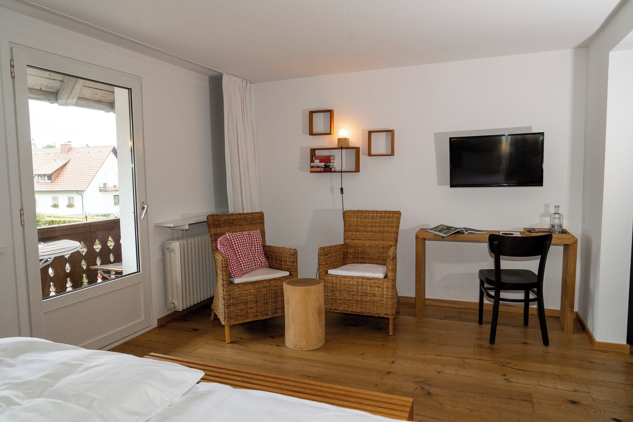Una habitación con una cama, dos sillones de mimbre, un taburete de madera, un escritorio con silla, un televisor montado en la pared y una ventana con vista a edificios.