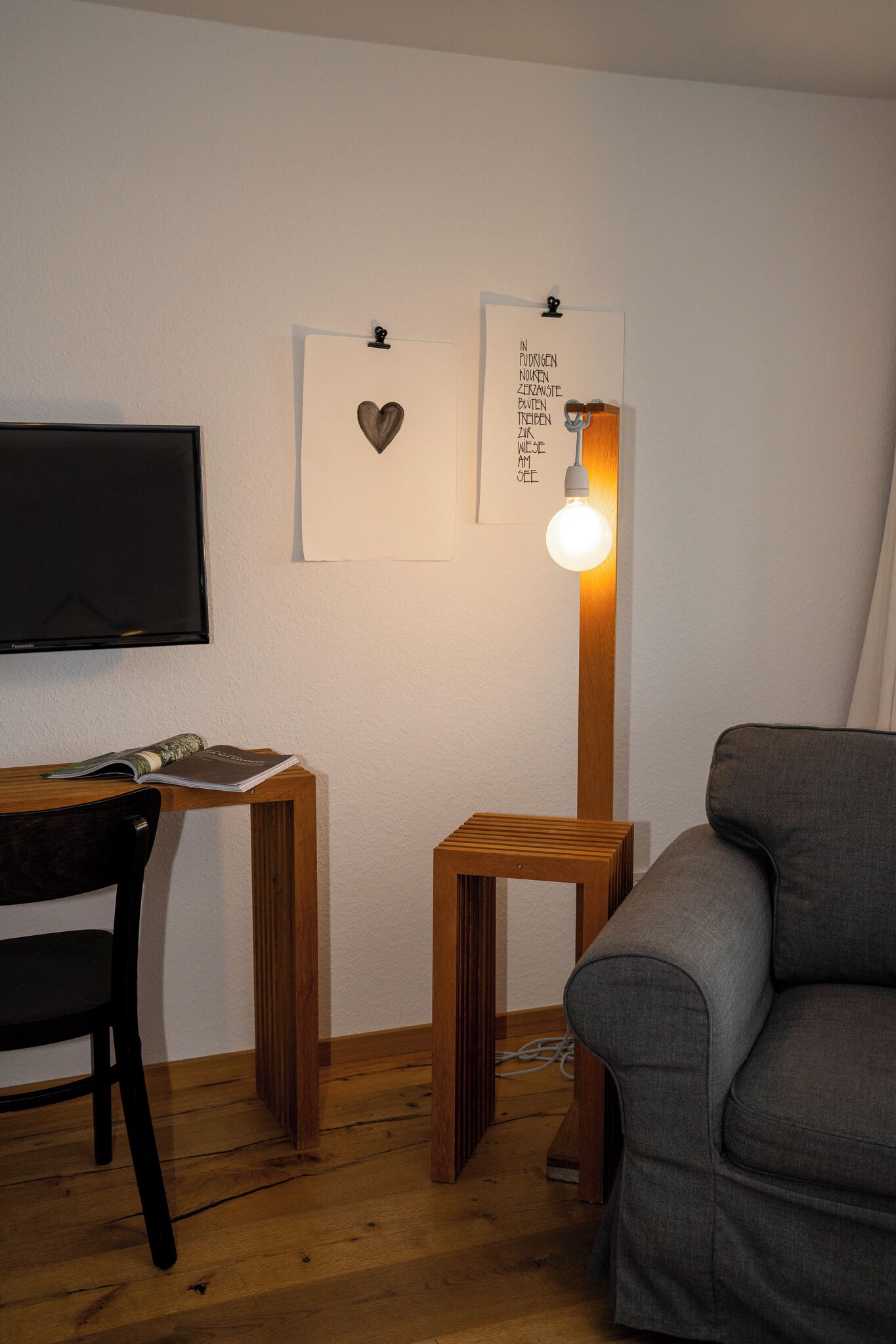Una habitación con un escritorio de madera, una silla, una mesita pequeña, un sofá gris y un televisor instalado en la pared.