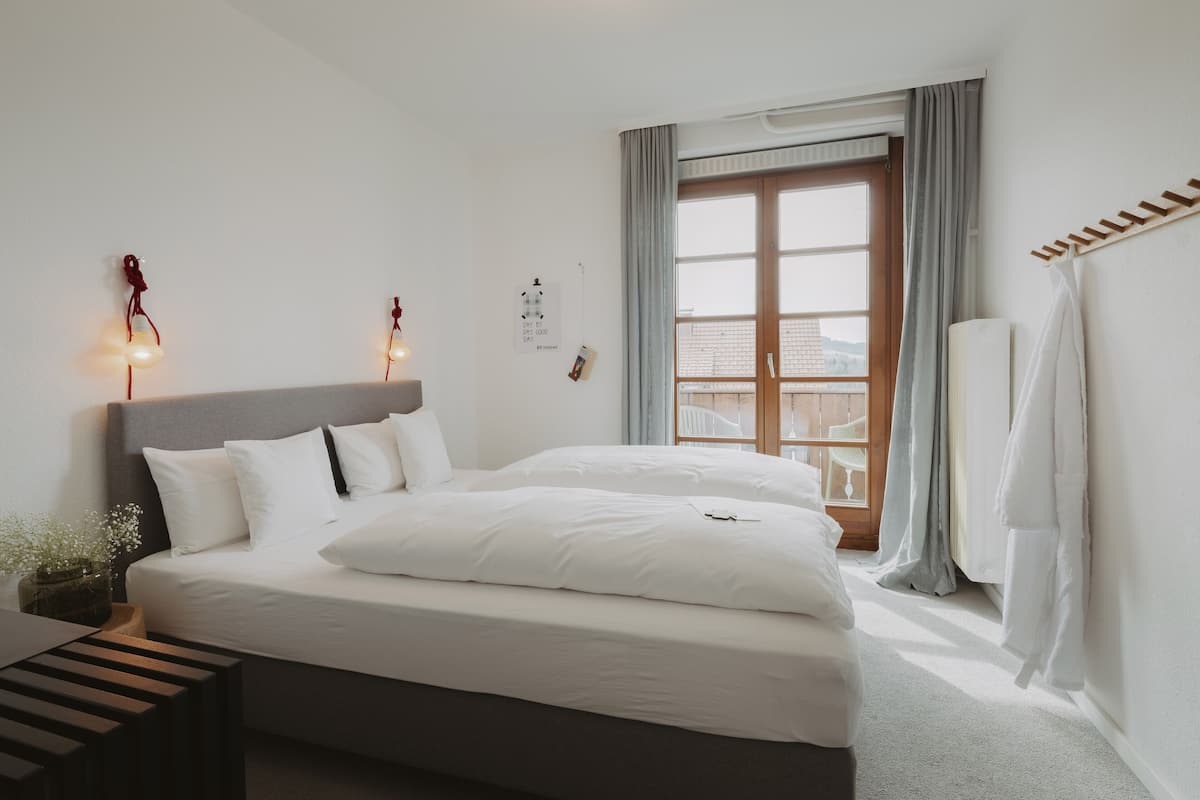 Una cama bien tendida con sábanas blancas, una mesita de noche con una lámpara y una ventana con cortinas.