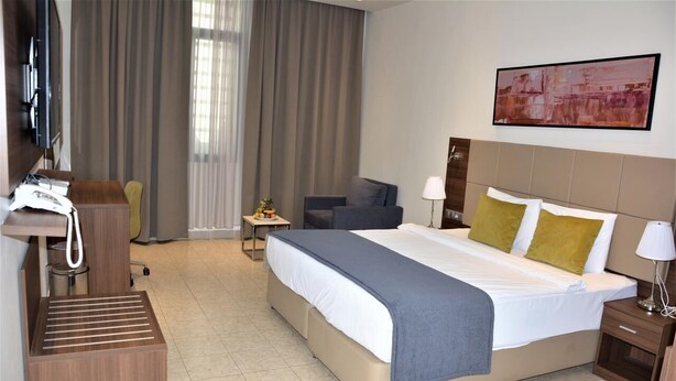 Ein Hotelzimmer mit einem Bett, einem Schreibtisch mit Telefon, einem Sessel, einem kleinen Tisch mit Lampe und einem Bild an der Wand.