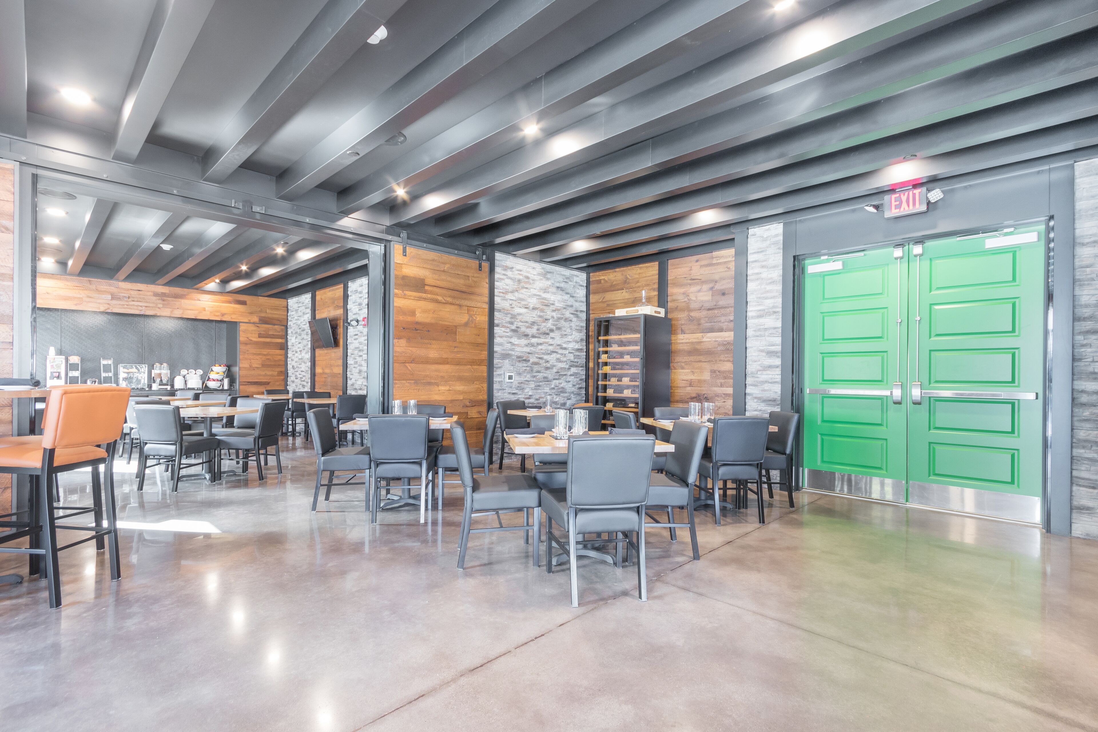 A modern dining area with tables and chairs, a green exit door, and a stone wall.
