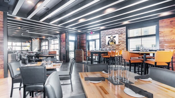 A modern restaurant with wooden tables and black leather chairs, featuring a brick wall and large windows. - Cobblestone Hotel and Suites Janesville (Janesville)