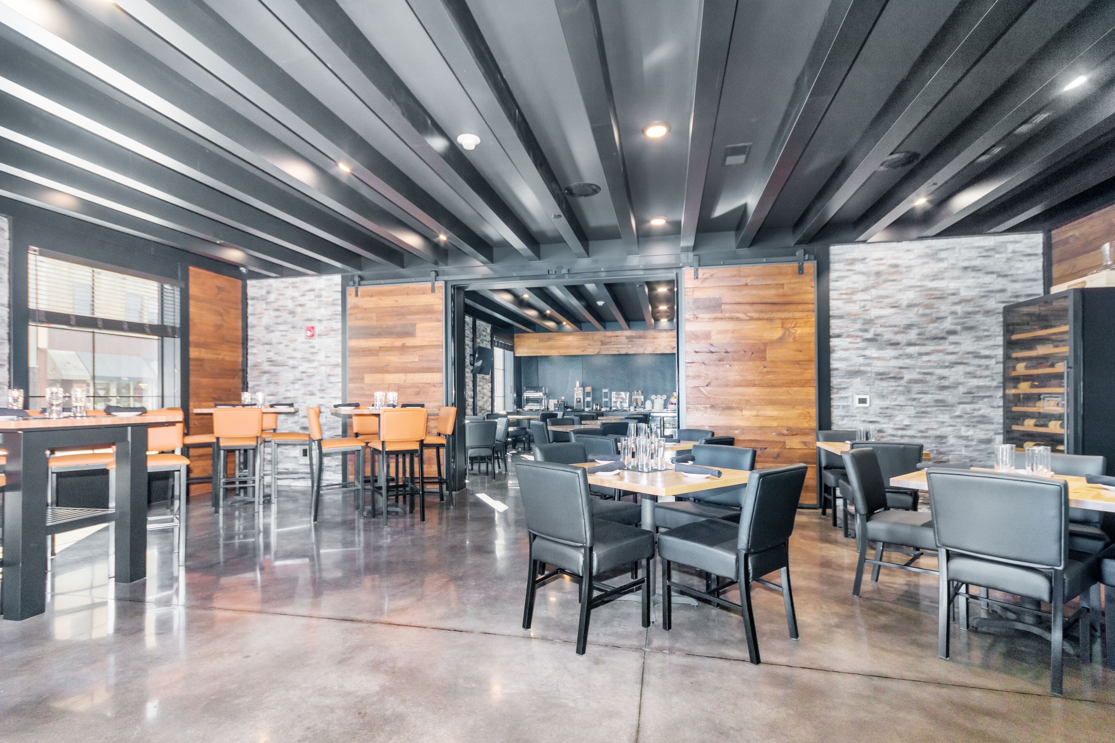 A modern dining area with wooden and stone walls, metal beams on the ceiling, and tables with chairs.