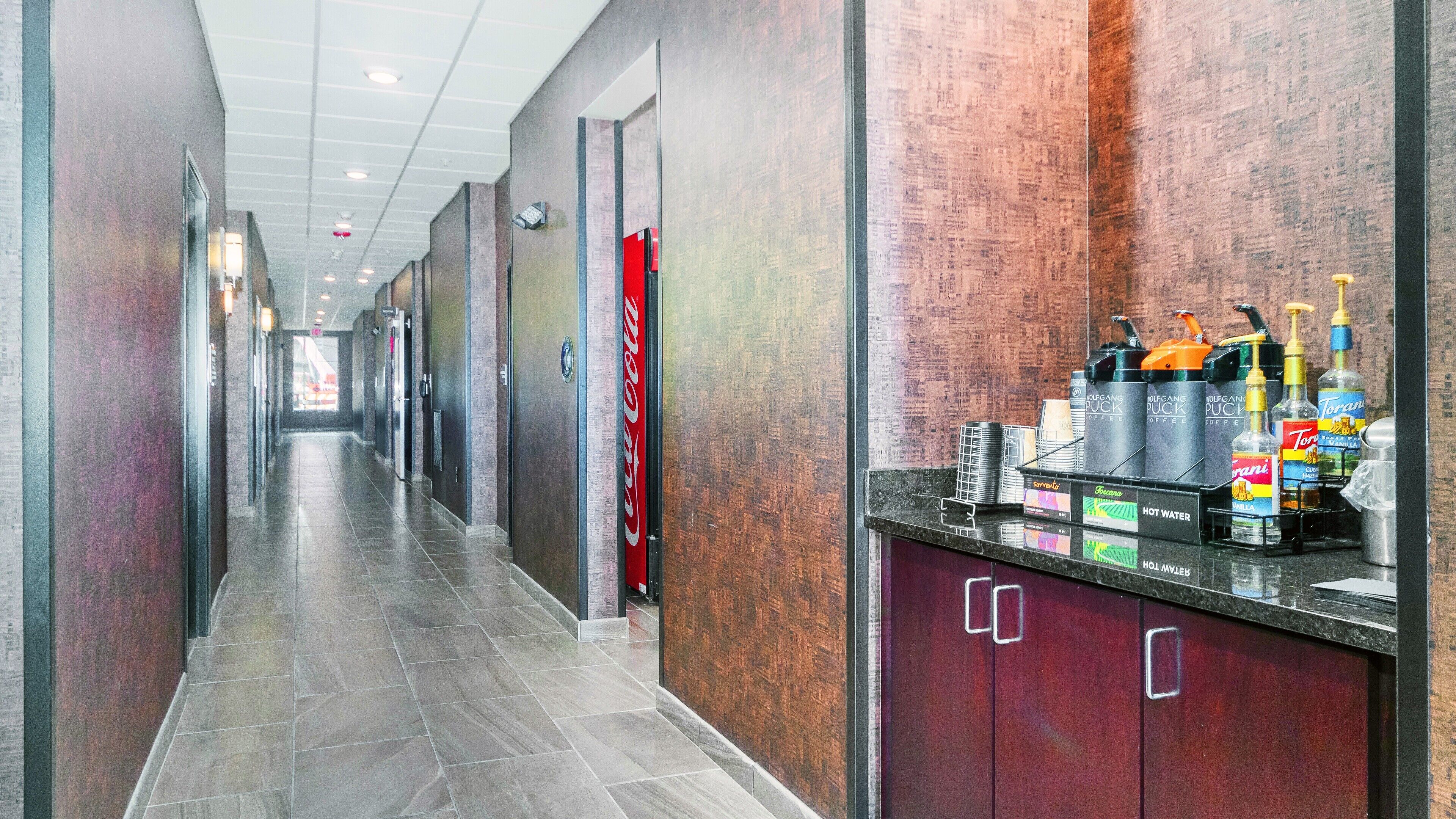 A hotel corridor with a reception area featuring a coffee machine, bottled water, and Torani syrup.
