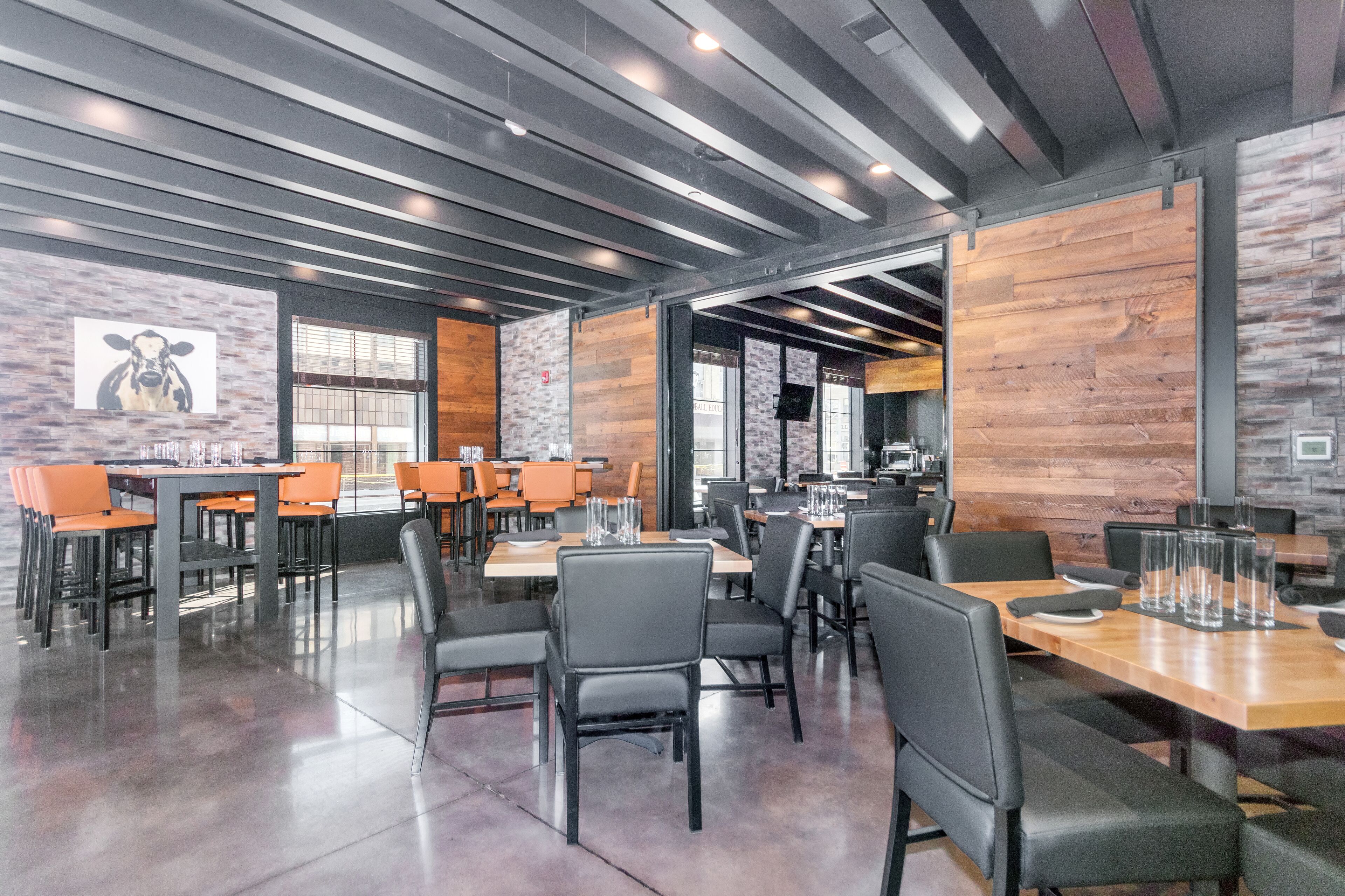 Un restaurante moderno con paredes de madera y piedra, sillas negras y anaranjadas, y un piso de concreto pulido.