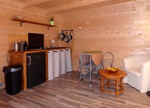 Eine kleine Küche mit Kühlschrank, Herd, Spüle und hängenden Töpfen. Im Wohnbereich befindet sich ein kleiner Esstisch mit Stühlen und ein weißes Sofa.