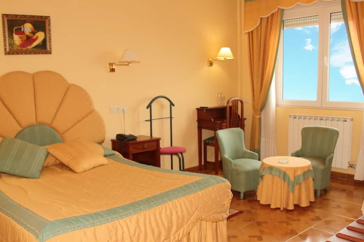 Een hotelkamer met een groot bed, twee fauteuils, een tafeltje, een bureau, een lamp en een raam met gordijnen.