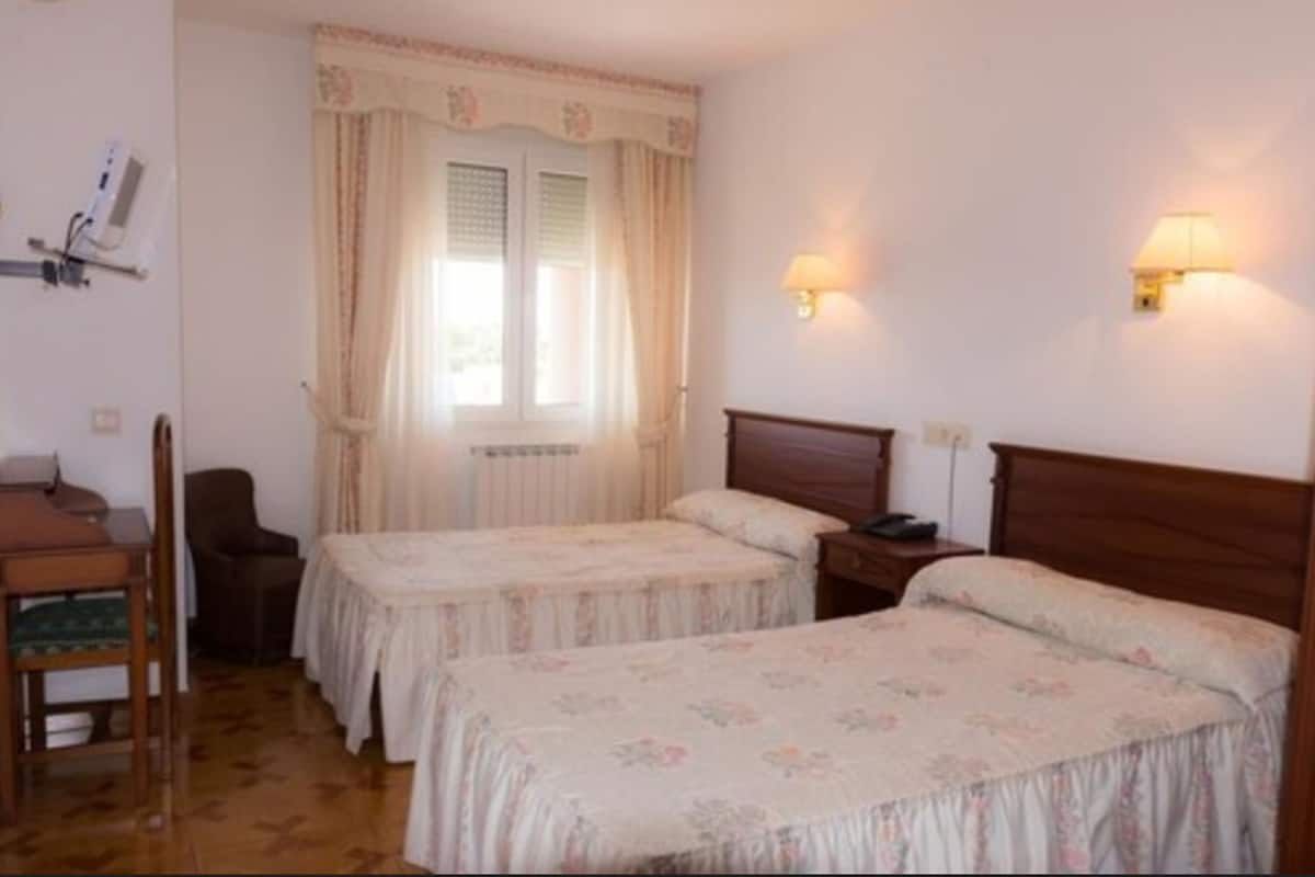 Een kamer met twee éénpersoonsbedden, een stoel, een bureau en een raam met gordijnen.