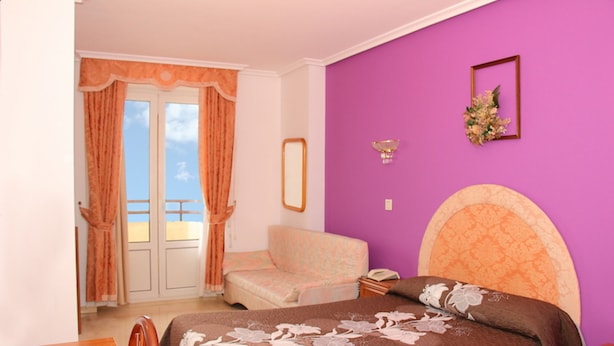 Een slaapkamer met een bed, een stoel, een raam met gordijnen, een spiegel aan de muur en een bloemenboeket.