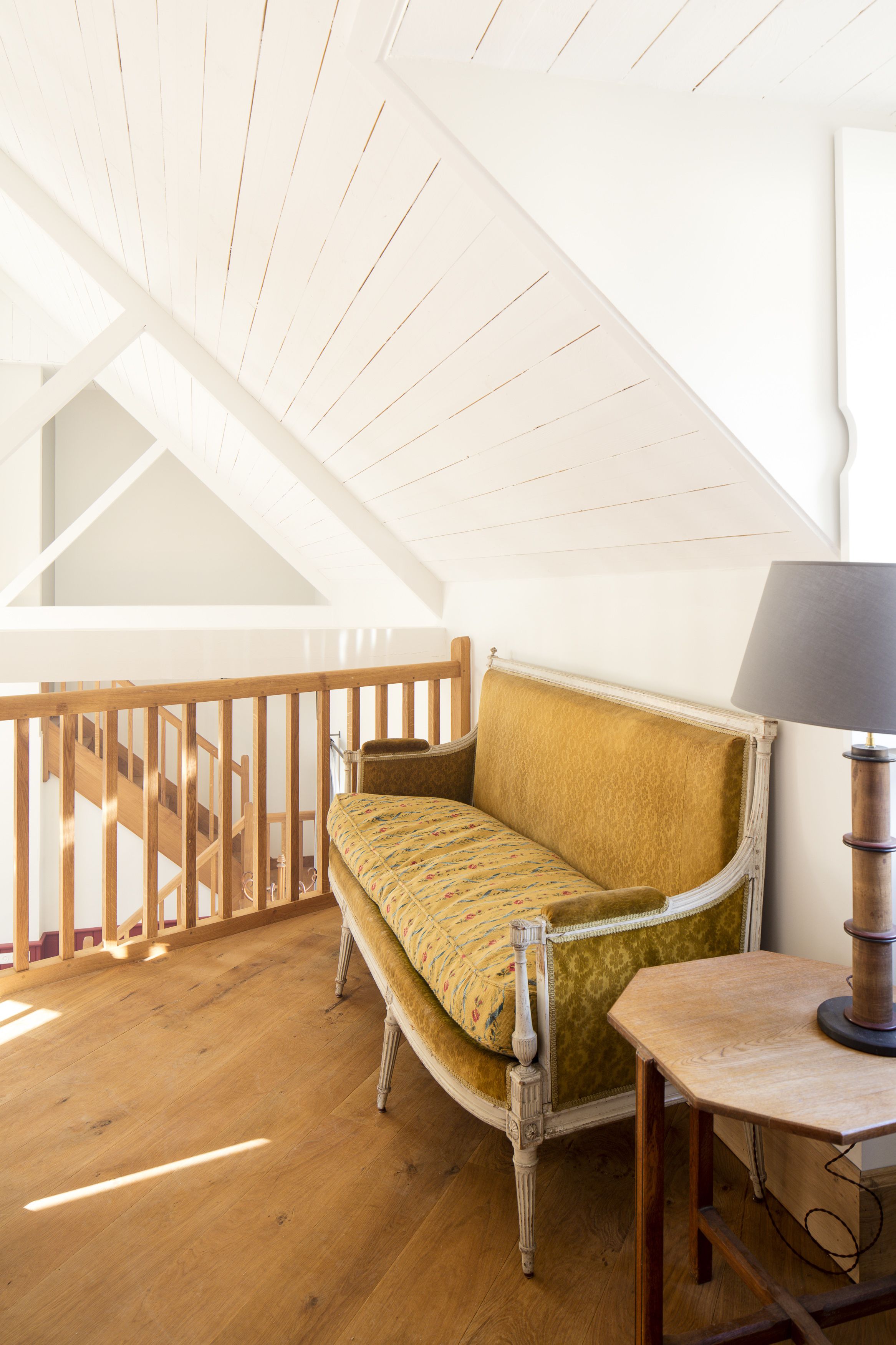 A high-ceilinged room with a wooden staircase, a vintage sofa, and a table with a lamp.