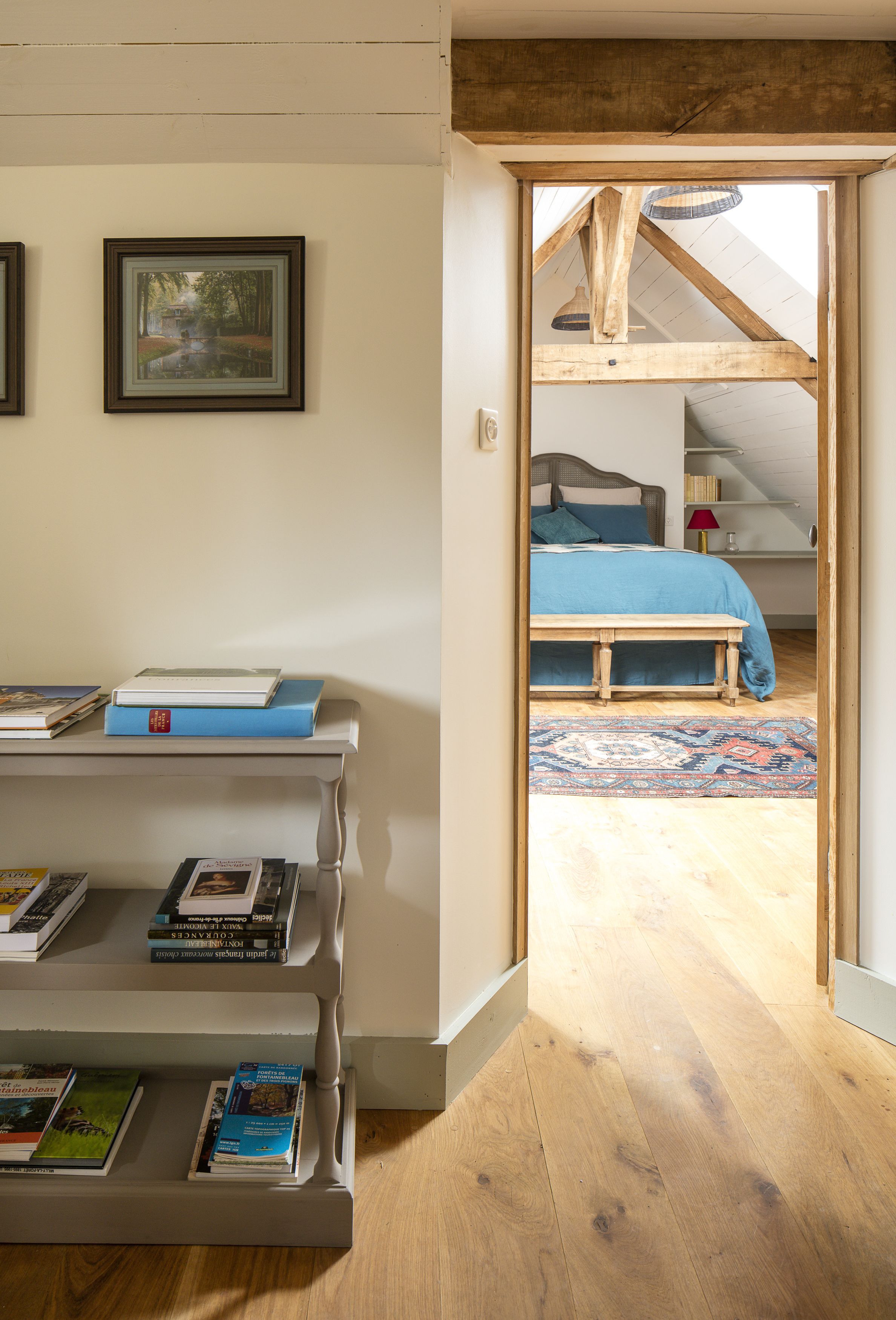 A room with a wooden floor, a bed with a blue bedspread, a wooden bench, and a bookshelf with books.
