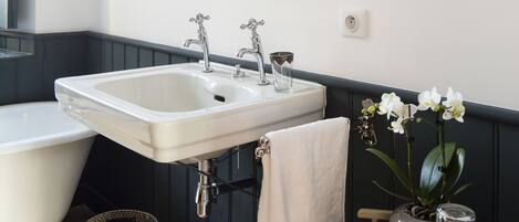 A bathroom with a white bathtub, a sink, and a mirror. There is a towel hanging on the wall and a small table with a vase of flowers.