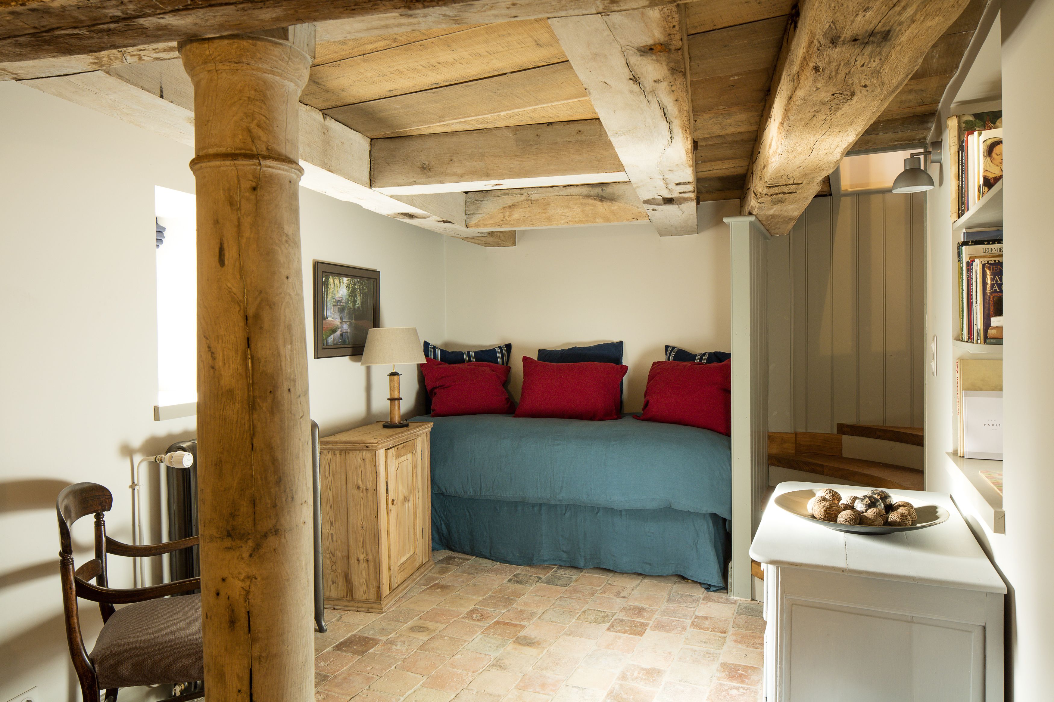 Una camera da letto con soffitto in legno, un letto con biancheria rossa e blu, un comodino con una lampada e una sedia.