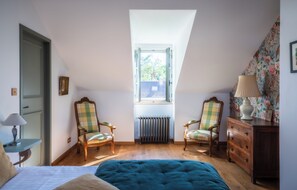 A bedroom with a sloped ceiling, two chairs, a dresser, a bed, and a window with a view. - Le Domaine de Courances (Courances)