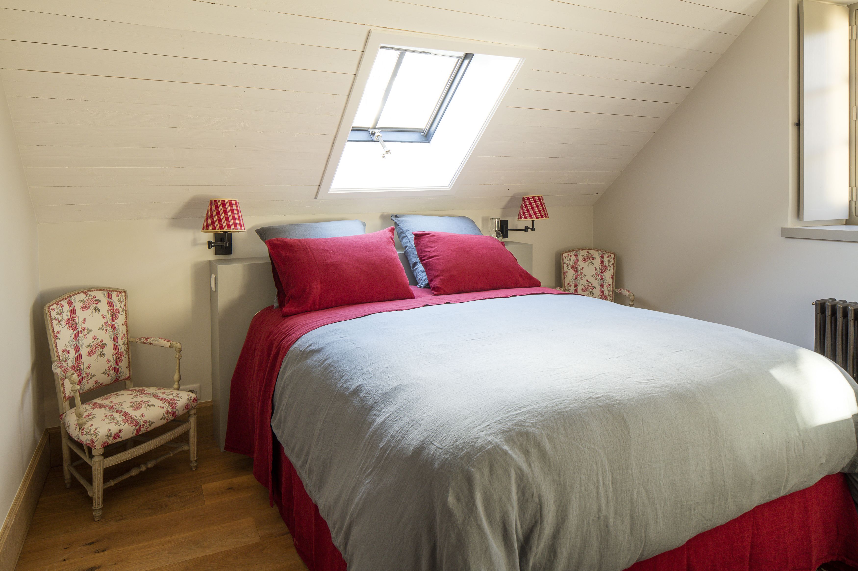 A bedroom with a sloped ceiling, a skylight, a bed with red bedding, two bedside lamps, a floral chair, and a radiator.