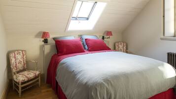A bedroom with a sloped ceiling, a skylight, a bed with red bedding, two bedside lamps, a floral chair, and a radiator.