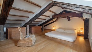 A cozy attic bedroom with a sloped ceiling, wooden beams, a bed with white linens, two wicker baskets, and two wall-mounted lamps. - The Lion House (Venice)