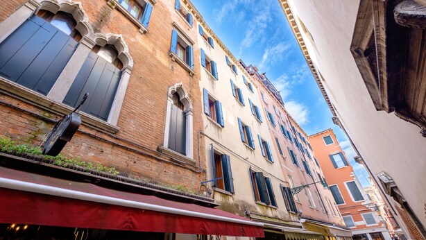 A narrow street with historic buildings, featuring arched windows and a red awning. - The Lion House (Venice)