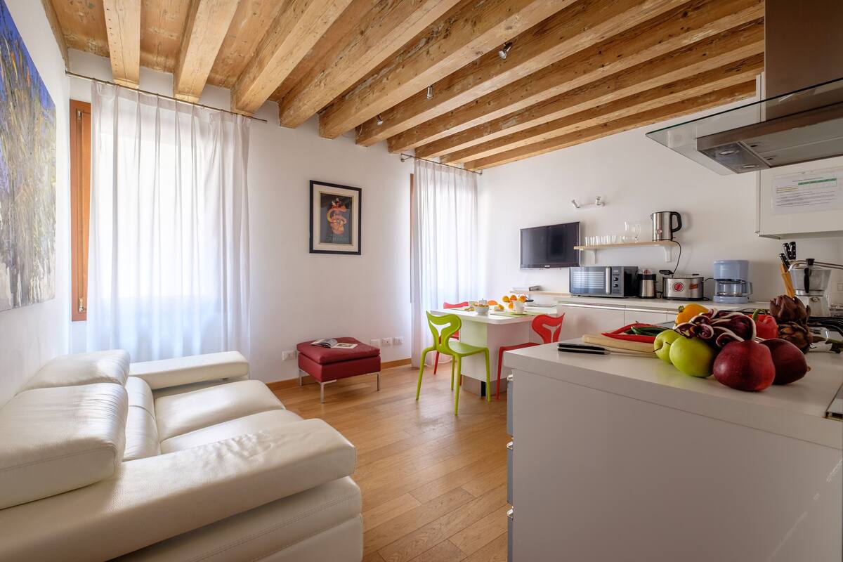 En moderne stue med sofasæt, et spiseområde med bord og stole, og et køkken med h årde hvidevarer og et udvalg af frugt.
