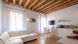 A modern living room with a white sofa, wooden ceiling beams, a dining area with a table and chairs, and a kitchen with appliances. - The Lion House (Venice)