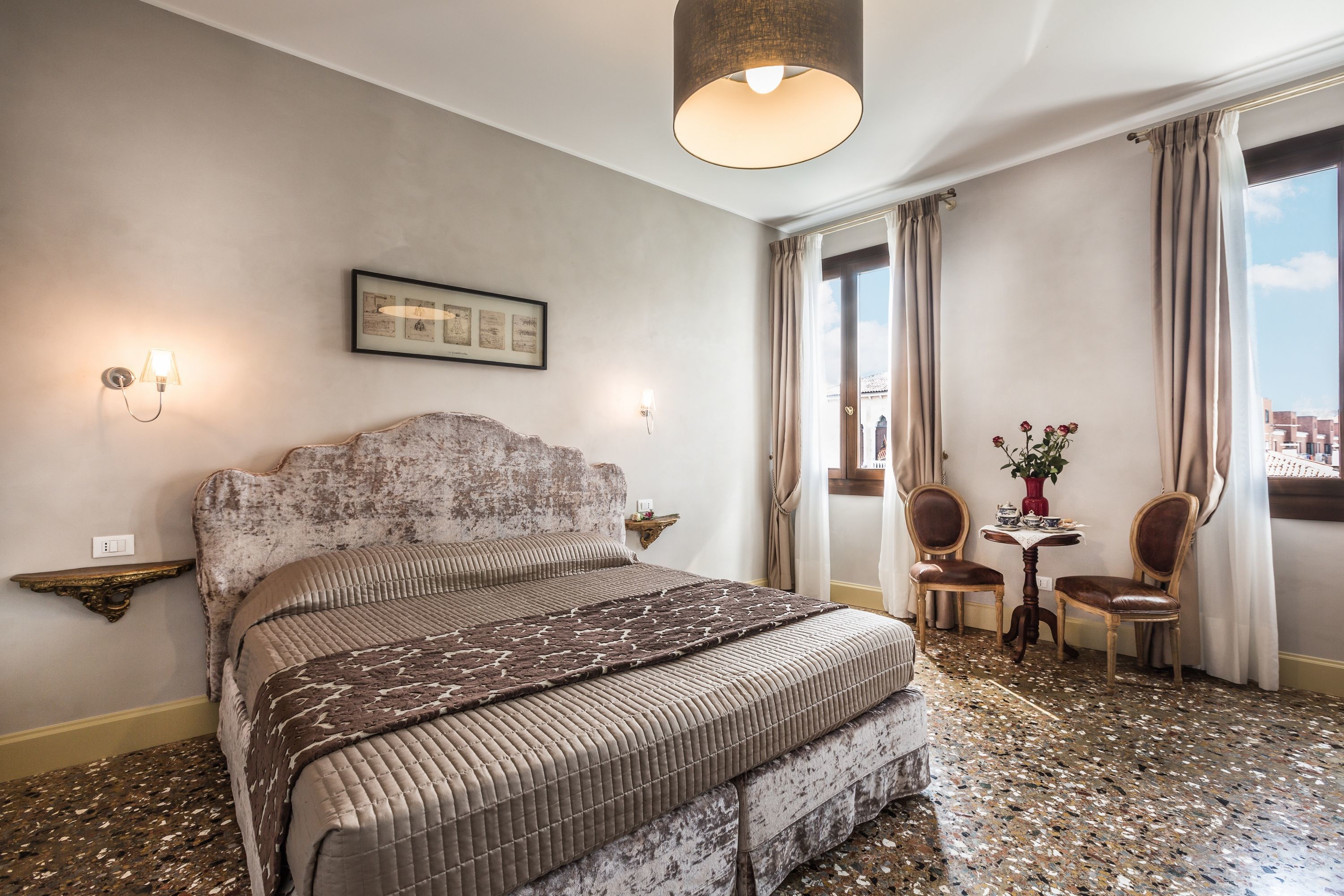 Une chambre d’hôtel avec un grand lit, une petite table avec des chaises, une étagère fixée au mur et une grande fenêtre avec des rideaux.