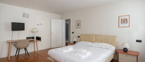 A bedroom with a bed, a desk, a chair, a TV, and a nightstand with a lamp.