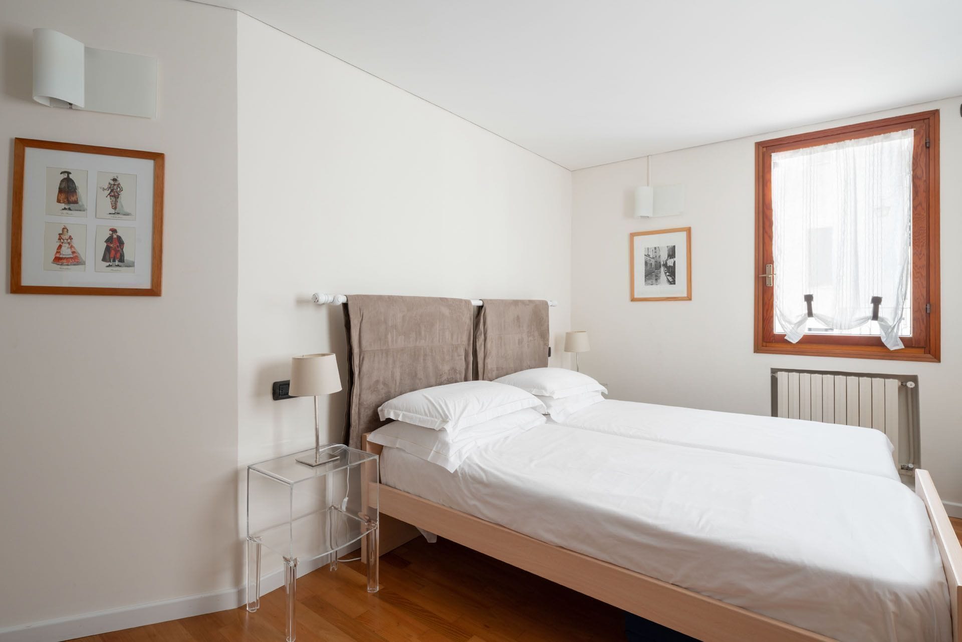Um quarto com cama, mesas de cabeceira, janela com cortinas e obra de arte na parede.