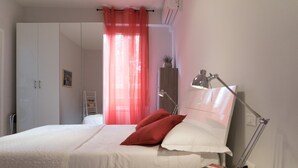 Ein Schlafzimmer mit einem Bett, einem Schreibtisch mit Lampe, einem Kleiderschrank und einem Fenster mit durchsichtigen Vorhängen.