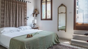 A bedroom with a bed, a green bedspread, a patterned rug, a window with curtains, a wall-mounted mirror, and a small table with a lamp. - Three Terraces (Venice)