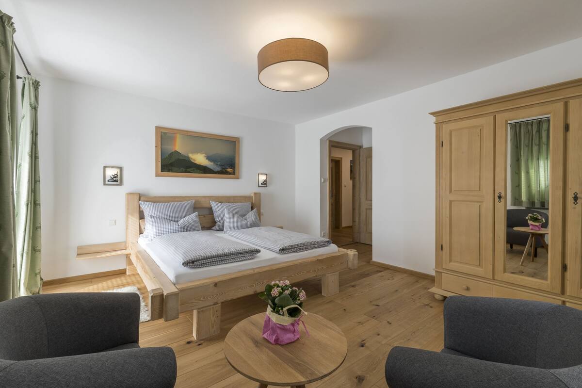 A bedroom with a bed, a round wooden table with a flower vase, two grey armchairs, a wardrobe, and a painting on the wall.