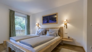 Quarto com cama de madeira, roupa de cama xadrez, duas luminárias na parede, um quadro e vista para o verde lá fora.