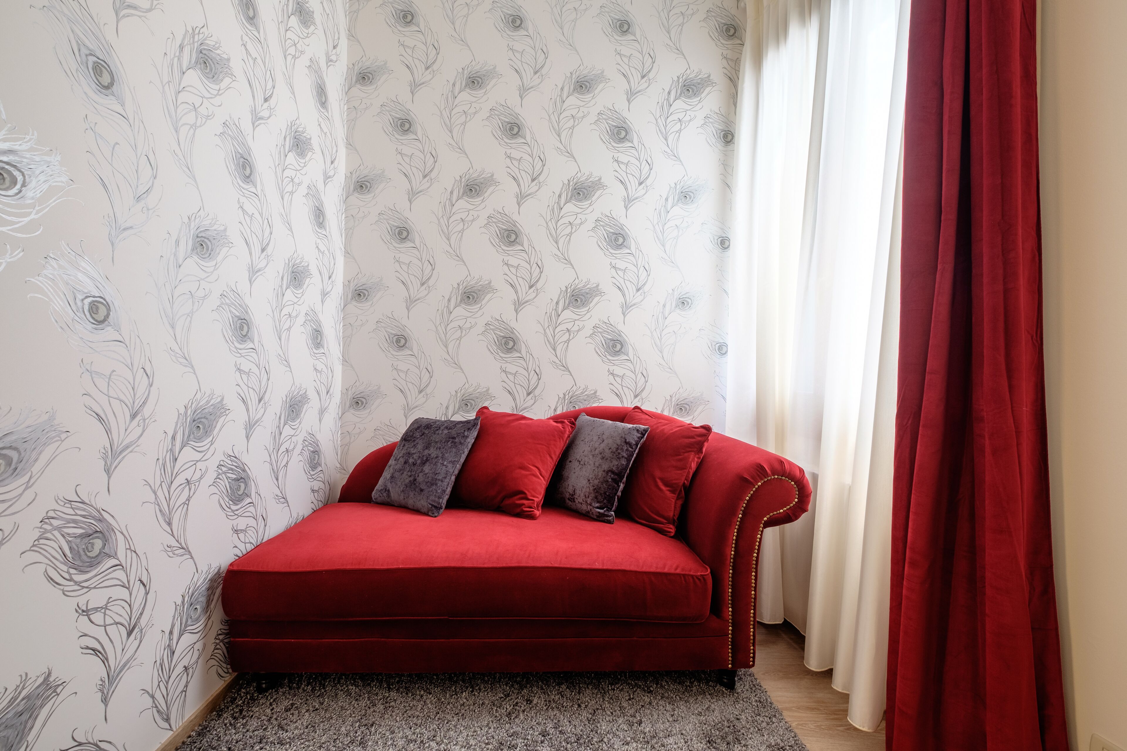 Un sofá rojo con cojines grises frente a una pared con estampado de plumas de pavo real.