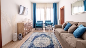 Ein Wohnzimmer mit einem beigen Sofa, zwei blauen Sesseln, einem Couchtisch, einem an der Wand montierten Fernseher, einer Holzkommode und einem gemusterten Teppich.