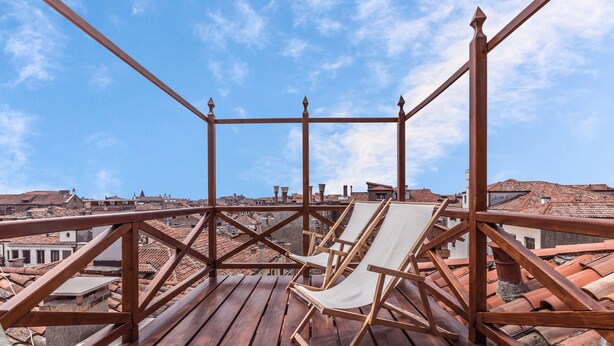 Eine Holzterrasse mit einem weißen Stuhl, mit Blick auf eine Stadtlandschaft mit Terrakotta-Dächern.
