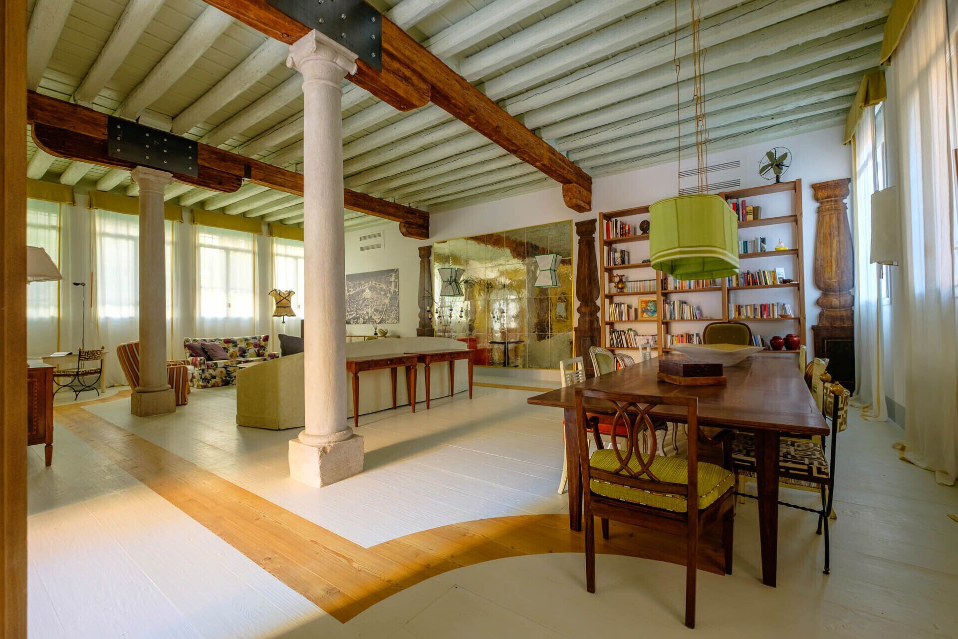 A spacious living area with a high ceiling, wooden beams, a large bookshelf, and a dining area with a wooden table and chairs.