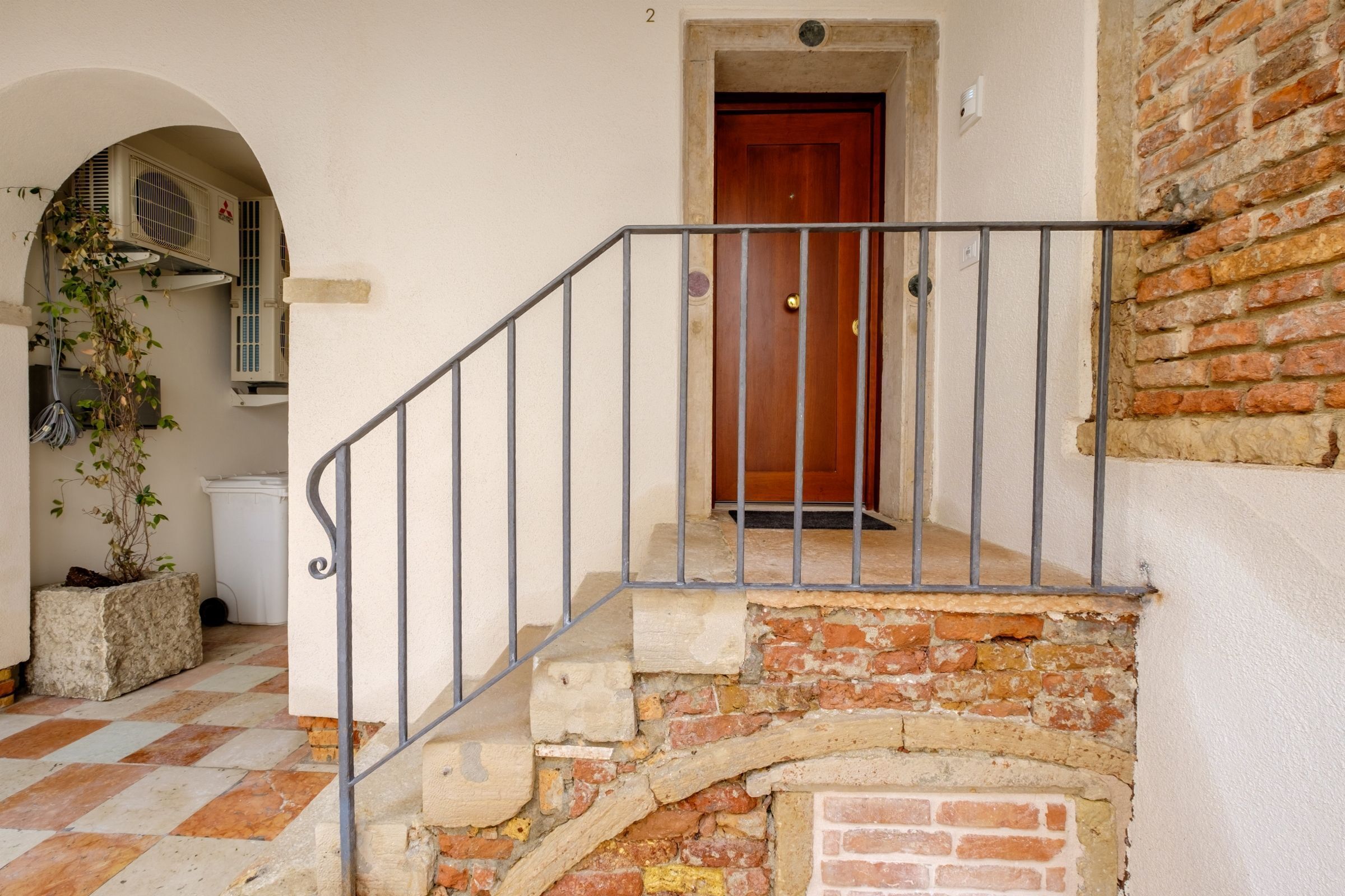 Una escalera de piedra que lleva a una puerta de madera, con barandilla de metal y una planta en maceta a la izquierda.