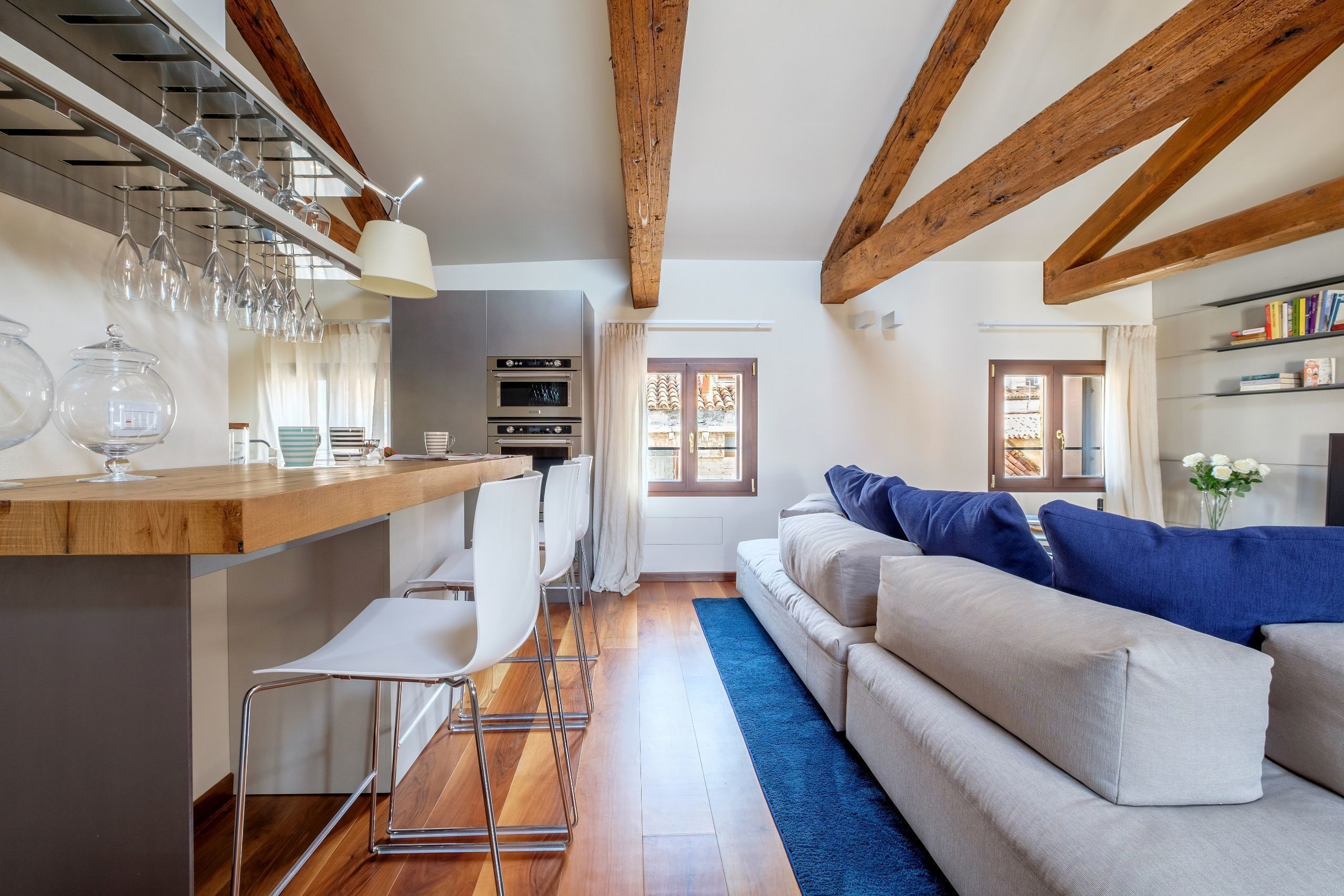 A modern kitchen with a wooden beam ceiling, a bar area with stools, and a sofa with blue cushions.
