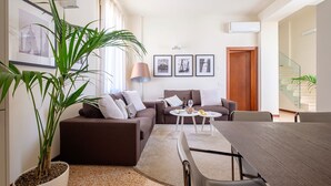 A modern living room with a brown sofa, a coffee table, and a dining table with chairs. There are three framed pictures on the wall, a potted plant, and an air conditioning unit. - Palazzo Ca' Moro apartments (Venice)