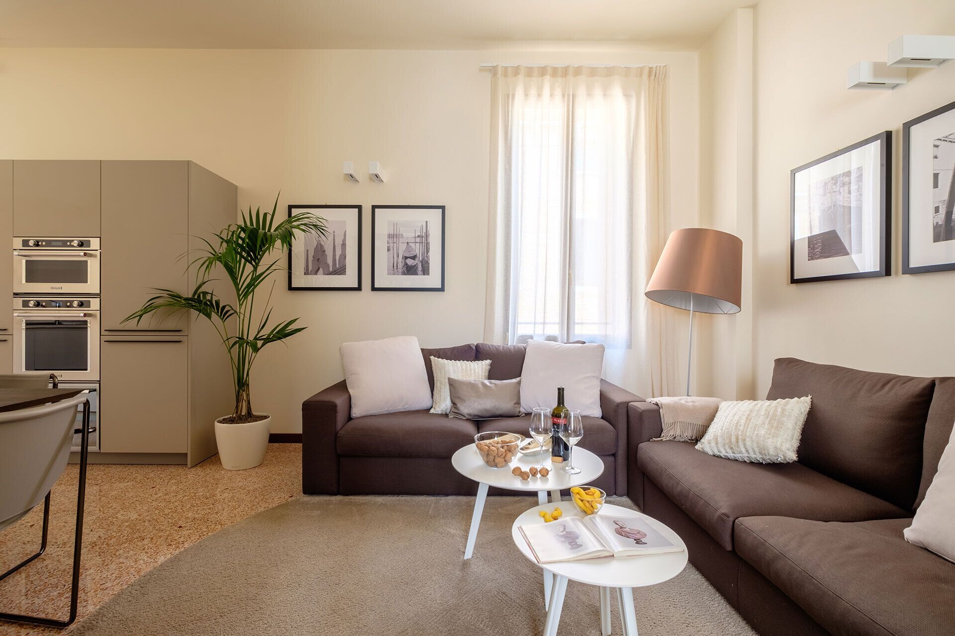 Un salon moderne avec un canapé marron, deux tables basses blanches, un coin repas avec une table et des chaises, une plante en pot et une grande fenêtre avec des rideaux.