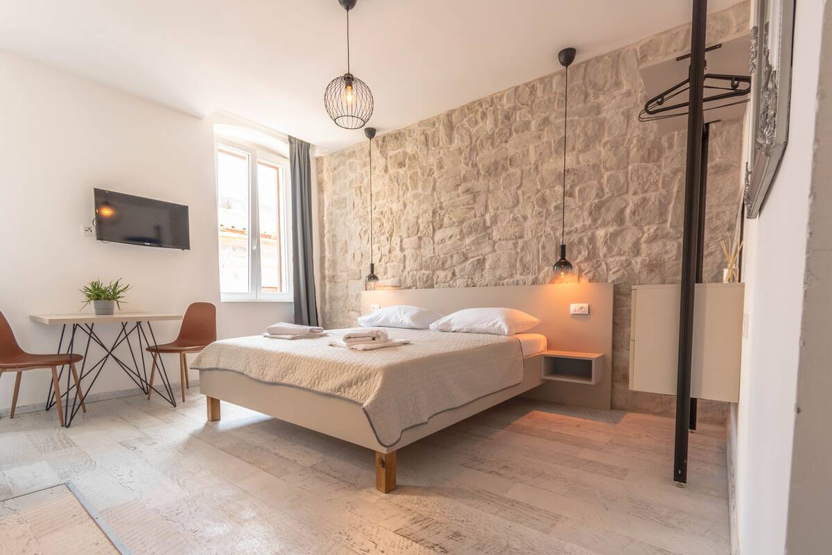 Een slaapkamer met een stenen muur, een bed met witte beddengoed, een bureau met twee stoelen, een televisie aan de muur en een hanglamp.