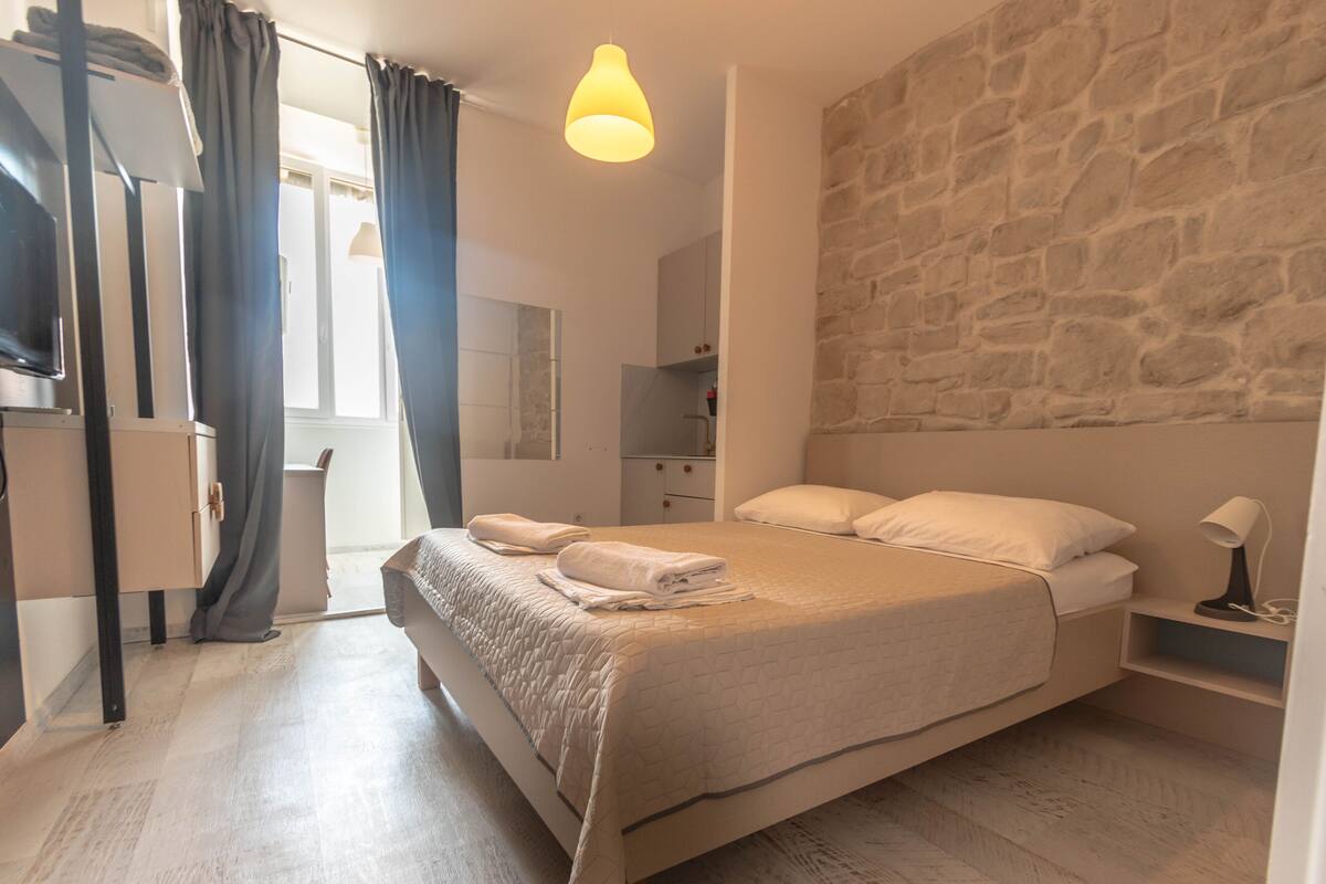 Een slaapkamer met een groot bed, een stenen muur, een nachtkastje met een lamp en een raam met gordijnen.
